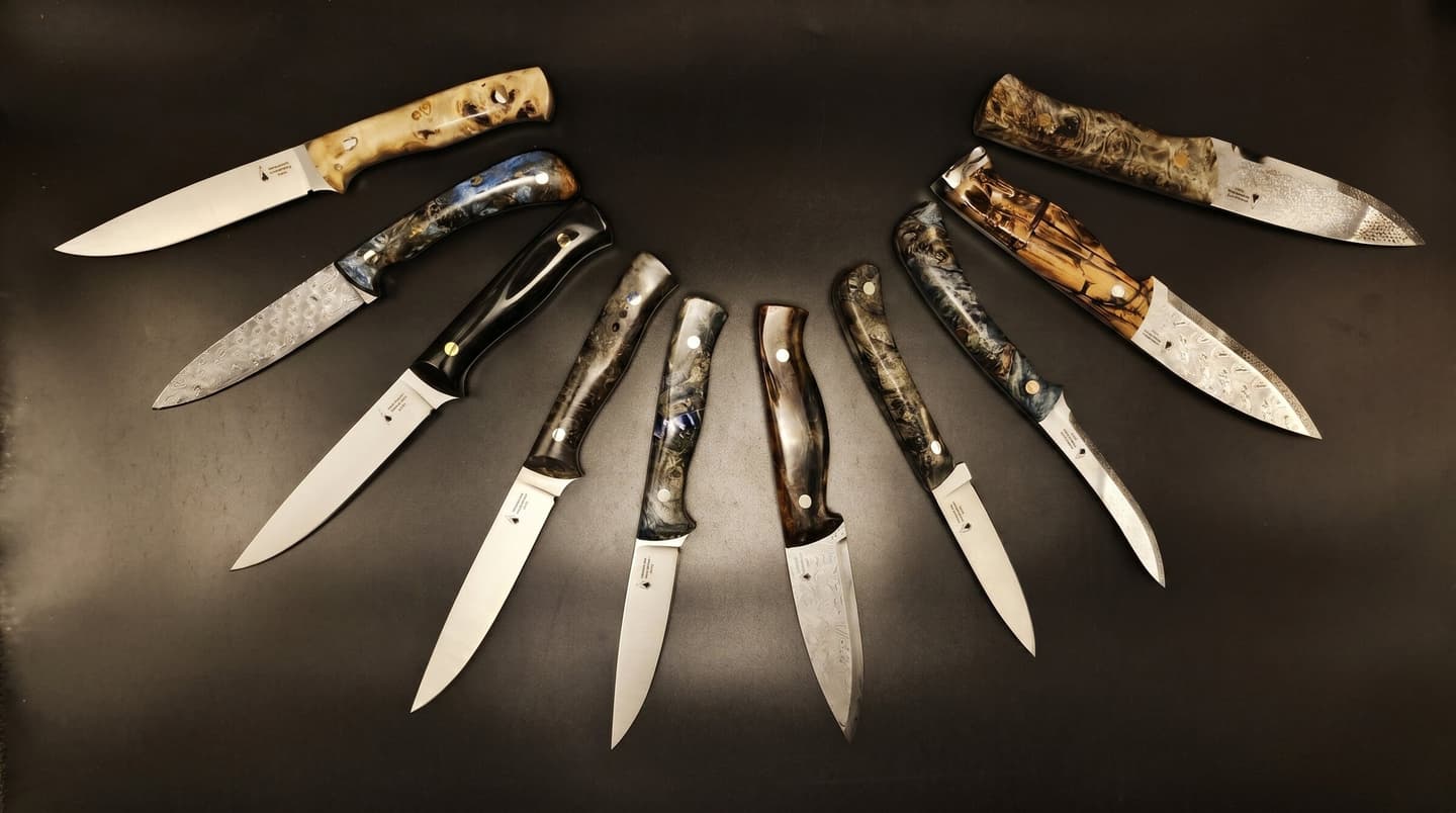 A wide fan of Emberleaf fixed-blade knives — Damascus and satin blades with exotic resin and burl handles — on a dark backdrop