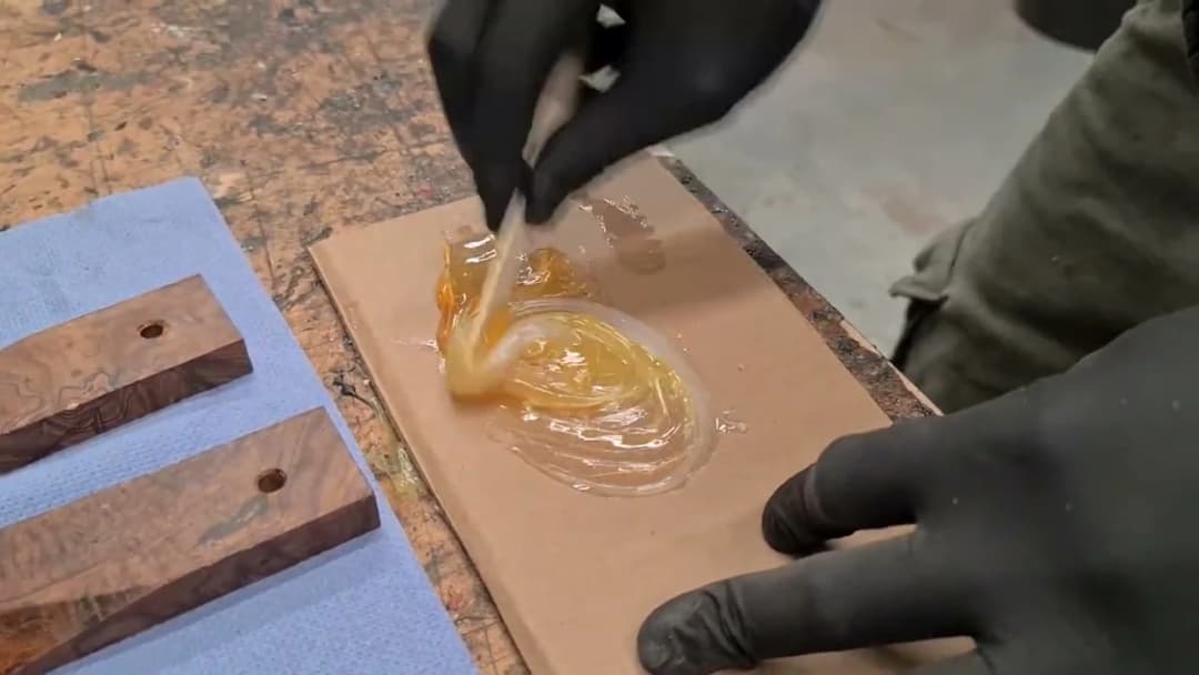 Black-gloved hands spreading two-part epoxy onto wooden handle scales beside the prepped tang